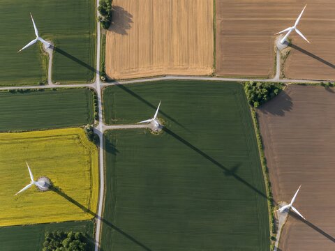 Top-down view of wind turbines casting long geometric shadows across cultivated farmland and narrow service roads.