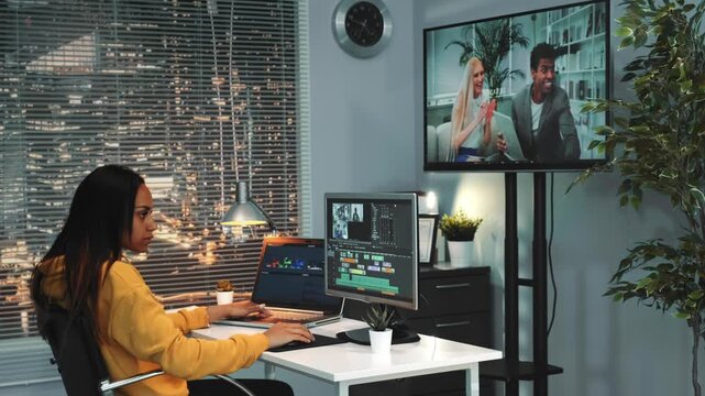 An African american woman with long dark hair edits video using multiple monitors in a professional nighttime office setting with city lights visible outside.