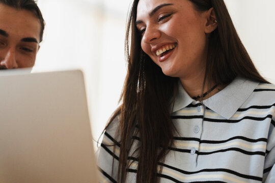A young man and woman look at a laptop computer together. They are smiling and laughing while watching something funny on the screen in a bright indoor room.
