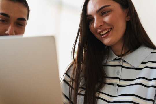 A young man and woman look at a laptop screen together. They are smiling while browsing content on the computer, sharing a pleasant moment in a bright indoor setting.