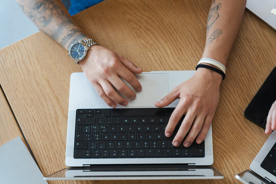 A tattooed person sits at a wooden desk and types on a modern laptop computer. Their tattooed hands rest on the keyboard in a top-down view of the workspace.