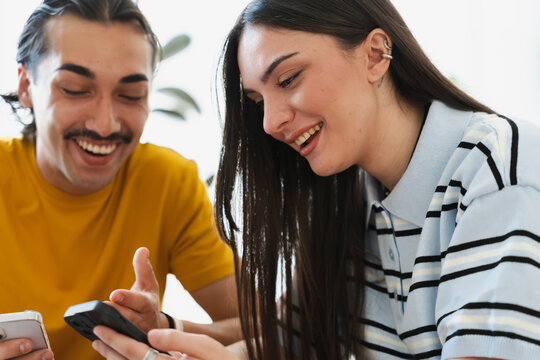 A young man and a woman are sitting indoors together, smiling and laughing happily while looking at a smartphone screen, sharing a cheerful and lighthearted moment.