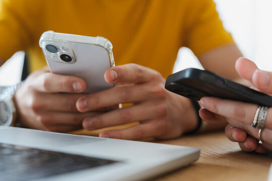 Two people sit together at a wooden table while using their smartphones. A laptop rests in the foreground as they browse devices and communicate digitally in an office.