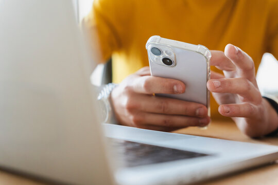 A person sitting at a desk wearing a yellow shirt is holding a modern smartphone in their hands. They are typing a message while working with a laptop computer in front of them.