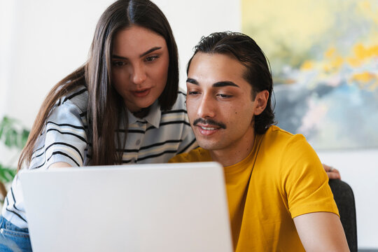 A young man and woman look at a laptop computer together indoors. The woman points at the screen while they collaborate on a project in their modern apartment living space.