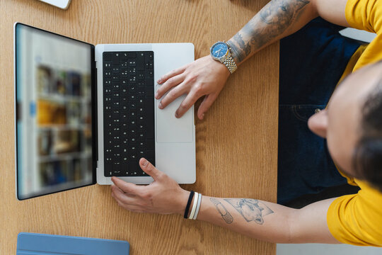 A tattooed man wearing a yellow shirt sits at a wooden table, browsing vacation rental listings and travel booking websites on his portable laptop computer.