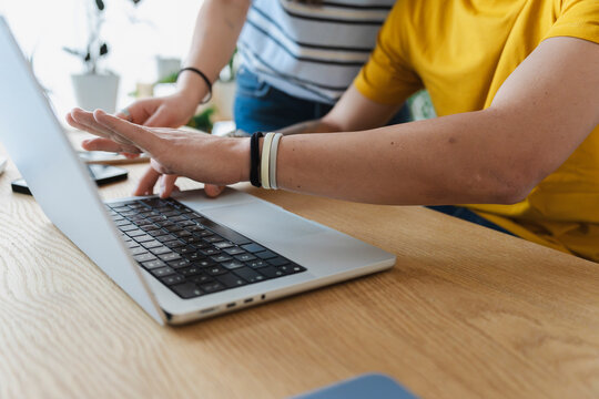 A young man and woman work together in a bright office, collaborating closely on a digital project while typing on a modern laptop computer placed on a wooden desk.