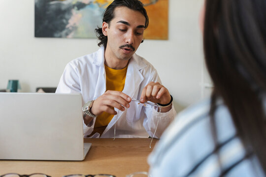 A male optician wearing a white coat sits at a desk with a laptop, holding up a pair of eyeglasses while talking to a female client during a professional consultation.