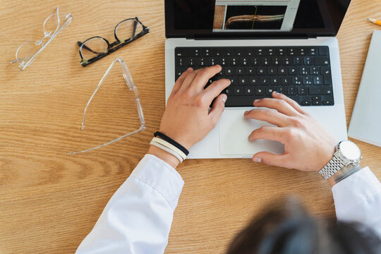 A doctor in a white coat sits at a wooden desk and types on a laptop. The computer screen displays a medical eye scan. Several pairs of eyeglasses rest nearby.