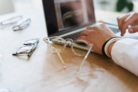 A person types on a laptop keyboard at a wooden desk. Transparent eyeglasses rest on the surface in the foreground, with other pairs scattered nearby in the bright workspace.