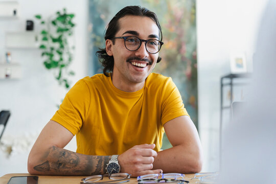A smiling young man with stylish glasses and a mustache sits at a wooden table inside a bright modern studio. He wears a yellow t-shirt and happily converses with someone.