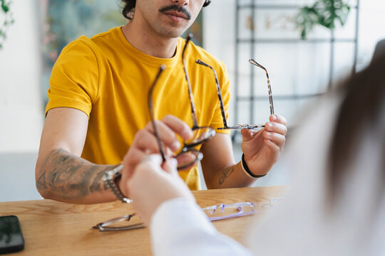 A young man wearing a yellow t-shirt sits at a wooden table in an optical store, holding up different pairs of eyeglass frames to choose his new glasses during a consultation.