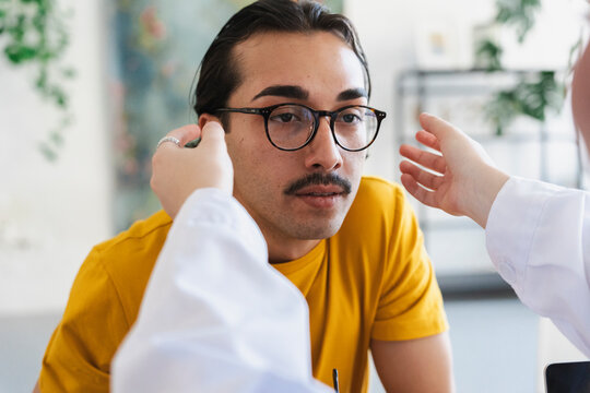 A young man wearing glasses sits in a medical office while a doctor examines his ears. The healthcare professional gently checks the patient during a routine clinical checkup.