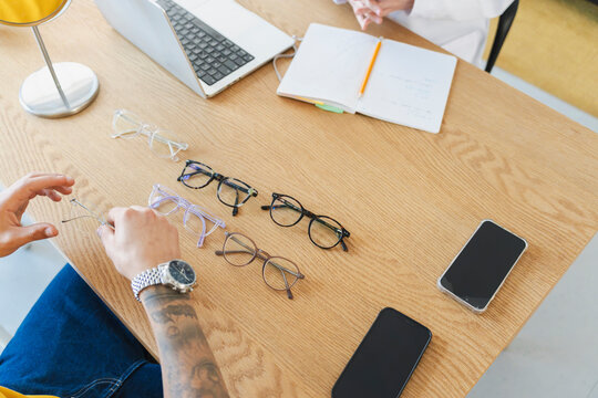 A customer with tattoos sits at a wooden table selecting new eyeglasses from several pairs displayed before them. An optician assists from across the desk with a laptop.