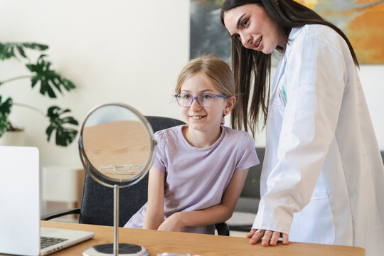 A young girl wearing new purple eyeglasses smiles while looking at her reflection in a mirror. A female optometrist in a white coat stands beside her during an eye exam.