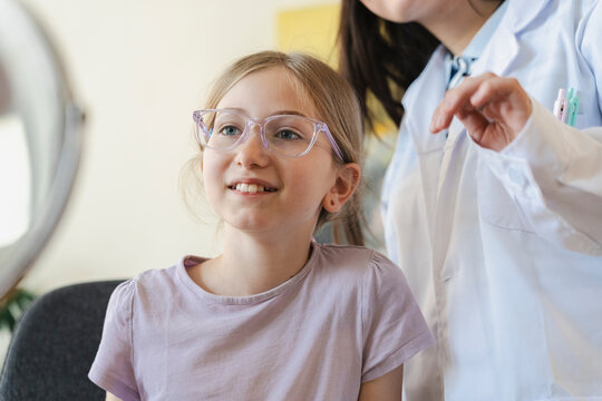 An optometrist in a white coat helps a young girl choose new glasses at an optical store. The smiling child tries on fashionable frames while testing her vision during the exam.