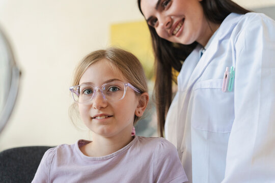 A young girl wearing new eyeglasses smiles while visiting an optometrist clinic. A friendly female doctor in a white coat stands behind her, providing expert medical care.