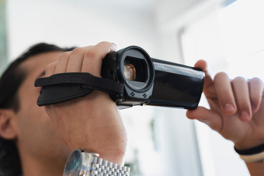 A young man holds a black digital video camera with both hands, carefully focusing the optical lens to record video footage indoors during bright daylight.