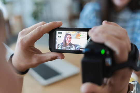 A person holds a digital video camera, recording a happy young woman whose smiling face is visible on the electronic viewfinder display during an indoor session.