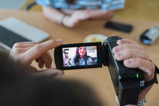 A person holds a digital video camera to record a young woman sitting at a table. The camcorder screen displays a clear live preview of her face during the indoor video shoot.