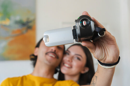 A happy young couple is recording a video selfie together in a bright room. The smiling man holds a compact digital camcorder up to capture the joyful moment.