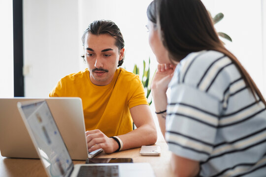 A young man wearing a yellow shirt and a woman with long hair sit together at a wooden table, working on laptops while collaborating on a business project.