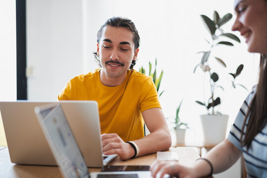 A young man with a mustache and his female colleague sit together at a wooden table in a bright office. They are focused on their laptops, collaborating on a work project.