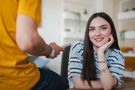 A young woman smiles happily while sitting at a desk indoors. A man stands beside her, holding up a smartphone to show her something interesting on the screen.