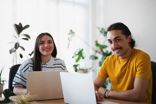 A young man and woman sit together at a wooden desk in a bright office, working on their laptops and smiling at the camera during a collaborative workday.