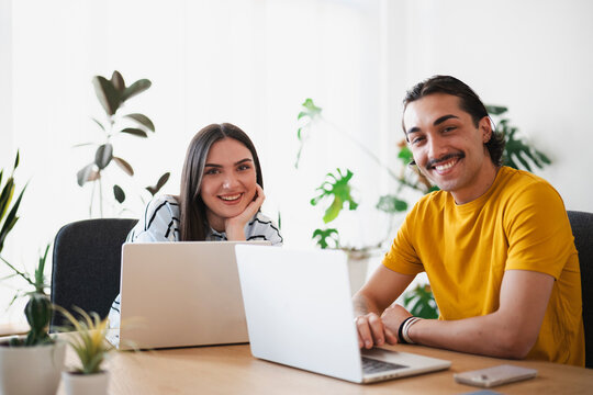 A young man and woman are working together on laptops in a bright, plant-filled office. They smile at the camera while collaborating on a modern project at their wooden desk.
