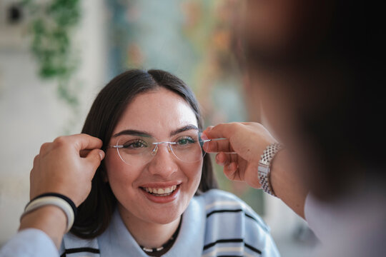 An optometrist helps a smiling young woman try on new rimless eyeglasses during her consultation at the store. She feels happy while testing the new frames.