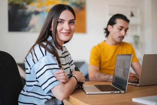 A young woman with dark hair sits at a desk with a laptop, smiling toward the camera. In the background, a man works on his computer in a bright modern home office.