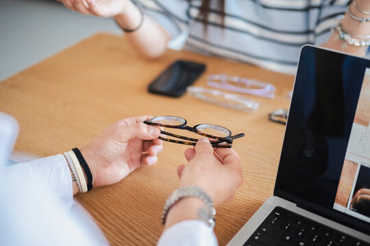 An optometrist holds a pair of eyeglasses during a consultation with a customer in an optical store. The professional discusses lens options at a wooden desk with a laptop.