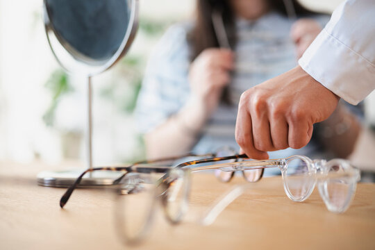 An optician selects a pair of eyeglasses from a wooden table while a customer sits in the background, preparing to try on different frames for a new vision prescription.