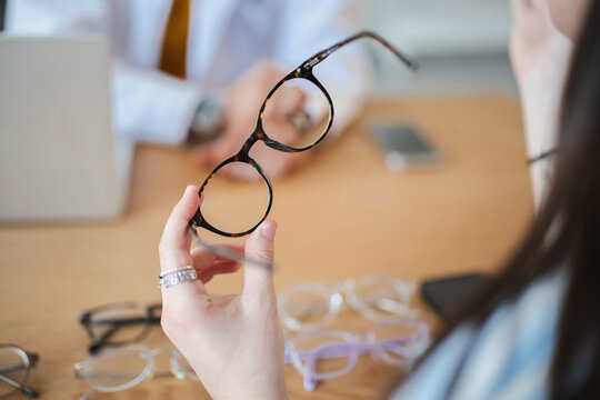 A woman sits at a wooden table in an optical store and examines a pair of stylish tortoiseshell eyeglasses. An optician waits in the background during her consultation.