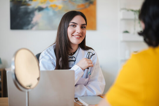 A friendly female doctor wearing a white lab coat sits at her desk during a medical consultation with a patient, smiling warmly at the camera inside a bright office.