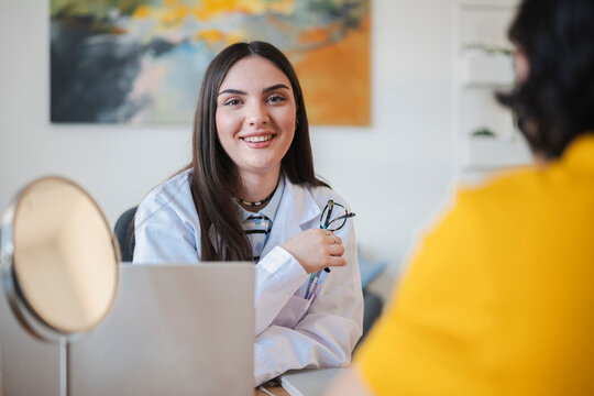 A smiling female doctor in a white lab coat sits at her desk during a medical consultation. She looks toward the camera while talking with a patient in a bright modern office.