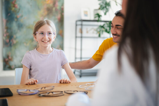 A young girl tries on new glasses at an optical store, smiling brightly. Her father sits beside her, supporting her during a consultation with an optometrist.