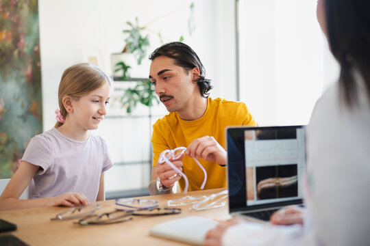 A father and his young daughter sit at a table in an optician's office, looking at various eyeglass frames together while choosing new glasses with the help of an optometrist.