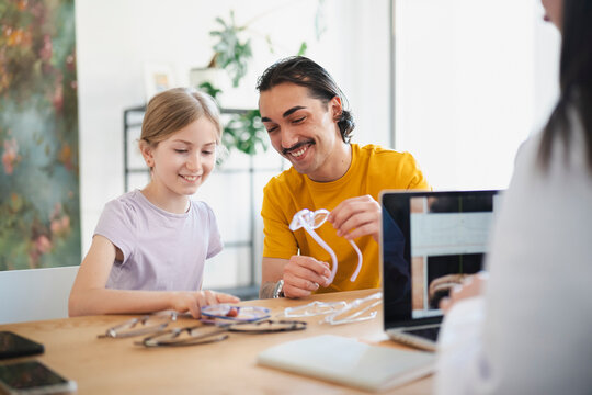 A young girl and her father sit together at an optician's office, smiling as they choose new eyeglasses from a table filled with various frames during an eye exam appointment.