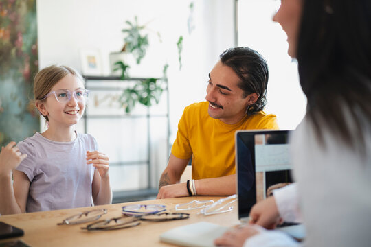 A young girl wearing new eyeglasses smiles at her father while sitting at a wooden table in an optical shop, trying on different frames with the help of an optometrist.