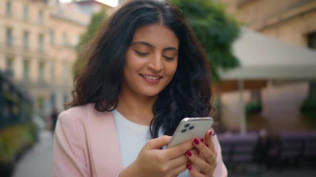 Happy smiling Indian woman walking in city street reading mobile phone outdoors Arabian ethnic girl female student businesswoman laughing toothy smile looking cellphone online chatting stroll outdoor