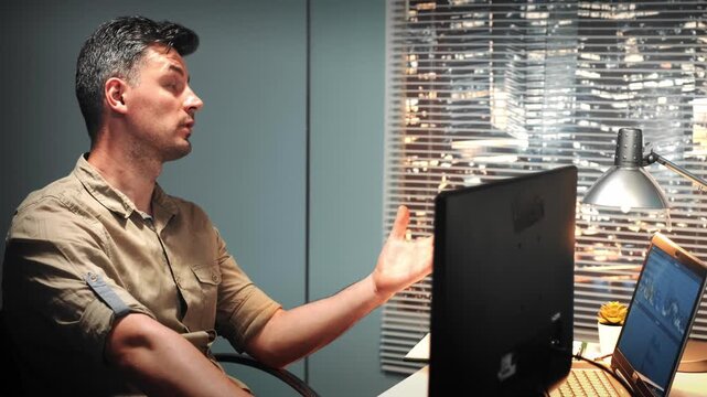A middle-aged man in a tan shirt is seated at a desk, gesturing while explaining a project in an office with multiple screens and a nighttime cityscape visible through a large window.