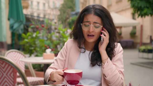 Happy smiling Indian woman talking on mobile phone drinking coffee in city cafe Arabian ethnic girl female businesswoman business call friendly talk smartphone discuss news visitor morning outdoors
