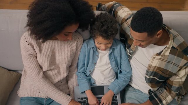 Top view African American family parents teaching little boy child typing laptop computer device ethnic mother father kid discussing online education inside living room couch learning digital school