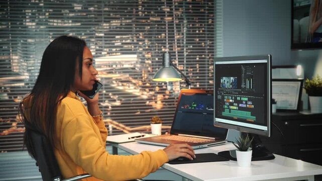A woman with long dark hair edits video on a laptop and speaks on a smartphone in a multimedia office at nighttime with city lights visible through a large window.