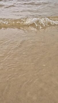 A sandy beach shoreline is shown close-up from above as gentle waves wash in and recede, creating foam bubbles that slowly dissolve over wet golden sand in bright daylight.