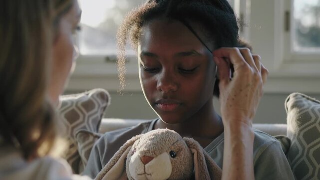 Reaching mom hand entering and smoothing child's curl, calming on couch by window with tan rabbit
