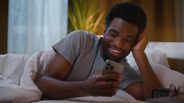 Happy African American man lying in bed holding mobile phone looking at smartphone screen watching video before sleep smiling ethnic guy male homeowner user relaxing at home bedroom late evening night