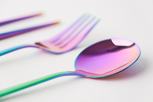 iridescent metallic sticks , fork and spoon resting on a glossy white surface, vibrant rainbow gradient reflecting soft pink and blue light